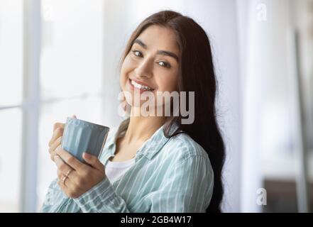 Porträt einer hübschen indischen Dame, die mit einer Tasse heißem Getränk in der Nähe des Fensters steht und die Kamera anlächelt Stockfoto