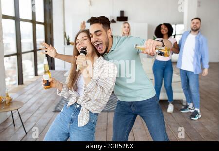 Tausendjähriges multiethnisches Paar, das Lied ins Mikrofon singt, Bier trinkt, mit Freunden zu Hause Party macht Stockfoto