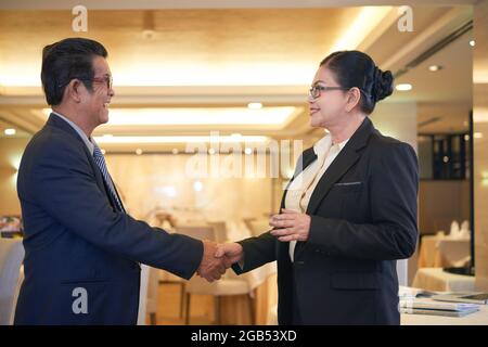 Lächelnder Senior Entrepreneur und seine weibliche Geschäftspartnerin schütteln sich die Hände und schauen sich gegenseitig an Stockfoto