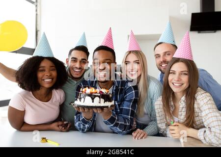 Porträt glücklicher multiethnischer Freunde, die Partyhüte tragen, mit Geburtstagskuchen posieren und drinnen die Kamera anlächeln Stockfoto