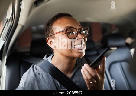 Afrikanische junge Frau in einer Brille, die während der Fahrt im Auto eine Audionachricht auf ihrem Mobiltelefon aufzeichnete Stockfoto