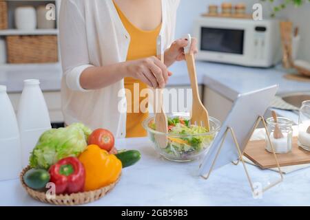Schöne lächelnde Frau essen Gemüse und gleichzeitig einen Salat aus einer Rezeptur auf der Tablet PC in einer Küche Stockfoto