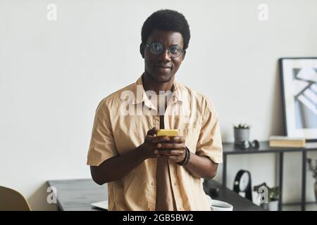 Porträt eines jungen afrikanischen Mannes, der im Büro auf die Kamera schaut und sein Mobiltelefon benutzt Stockfoto