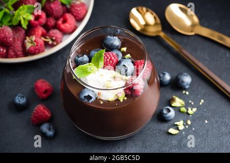 Dunkle Schokoladenmousse mit Pistazien, Beeren und Creme im Glas, schwarzer Steinhintergrund, Nahaufnahme Stockfoto