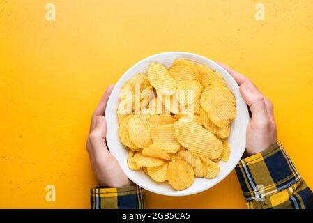 Kartoffelchips oder Chips in der Schüssel in männlichen Händen auf gelbem Hintergrund, Draufsicht, Kopierraum. Superbowl Snack, Junk Food Stockfoto