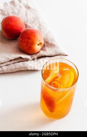 Ein Glas hausgemachter kalter Pfirsich-Eistee mit frischen Pfirsichen auf der Bettwäsche im Hintergrund. Stockfoto