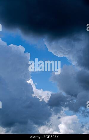 Lücke in weißen und grauen Cumuluswolken, die an einem regnerischen Sommertag einen blauen Himmel zeigen Stockfoto