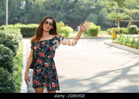 Lächelnde junge Frau in Blumenkleid und Sonnenbrille, die ein Taxi nimmt Stockfoto