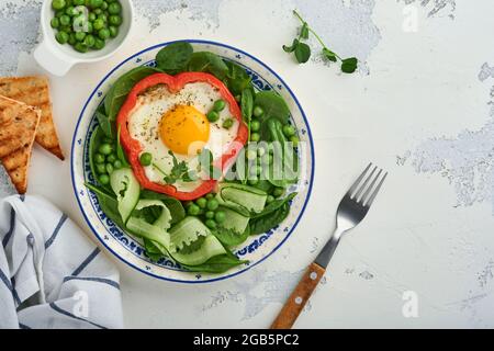 Rote Paprika, gefüllt mit Eiern, Spinatblättern, grünen Erbsen und Mikrogemüse auf einem Frühstücksteller auf hellgrauem Tischhintergrund. Draufsicht. Stockfoto