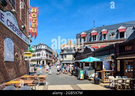 Touristen, die in der Monge-Straße in Etretat, Frankreich, spazieren, gesäumt von Straßencafés, Restaurants, Eisdielen und Hotels an einem sonnigen Tag. Stockfoto