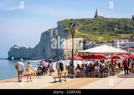 Menschen, die einen Drink in einem Strandcafé an der Küste in Etretat, Frankreich, mit der Kapelle Notre-Dame de la Garde mit Blick auf die Klippe des Amont und seinen Bogen genießen. Stockfoto