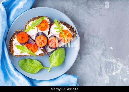 Köstliche geröstete Sandwiches mit Frischkäse, Kirschtomaten und Radieschen auf grauem Teller. Sandwiches mit Frischkäse und frischen Kräutern auf grauem Backgro Stockfoto
