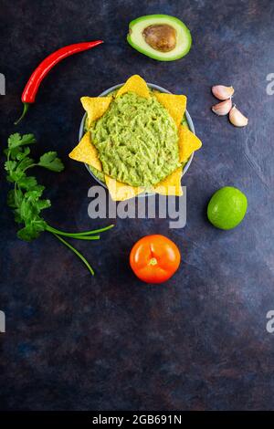 Guacamole mit Zutaten und Nachos-Tortilla-Chips. Avocado Guacamole mit Zutaten wie Pfeffer, Limette und Mais-Nachos auf dunklem Grund. Speicherplatz kopieren Stockfoto