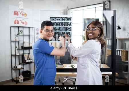 Konzept für Gesundheitswesen, Medizin und Radiologie. Zwei multiethnische Ärzte, eine afrikanische Dame und ein arabischer Mann, zeigten auf einen Röntgenscan und lächelten auf die Kamera in einem modernen, hellen Büro in der Klinik. Stockfoto