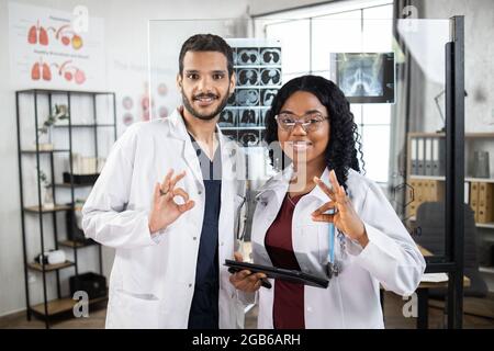 Porträt von zwei multikulturellen Ärzten in weißen Laborkittel, die im Krankenhauszimmer posieren und ein Schild mit der Kamera zeigen. Konzept der Medizin, Zusammenarbeit und Gesundheitsversorgung. Stockfoto