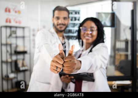 Verschwommene Ansicht von zwei fröhlichen multirassischen Ärzten in weißen Laborkittel, die Daumen nach oben zeigen, während sie im medizinischen Zentrum zusammen stehen. Konzept von Menschen, Medizin und Teamarbeit. Konzentrieren Sie sich auf die Hände Stockfoto