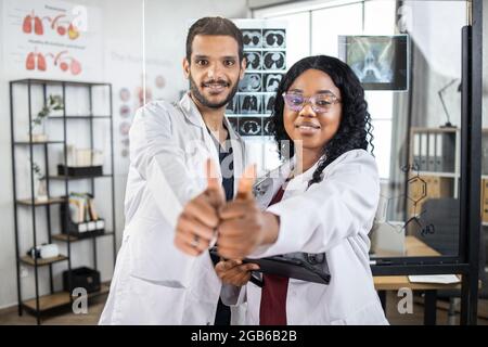 Zwei selbstbewusste medizinische Wissenschaftler stehen am Sitzungssaal zusammen, lächeln und blicken auf die Kamera und zeigen den Daumen nach oben. Zwei verschiedene Ärzte, Mann und Frau, posieren nach erfolgreichem Treffen in Innenräumen. Stockfoto