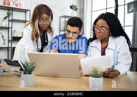 Drei fokussierte multiethnische Ärzte, die eine Online-Konferenz im Sitzungssaal des Krankenhauses abhalten. Medizinische Mitarbeiter und -innen, die während des Meetings einen modernen Laptop verwenden. Stockfoto