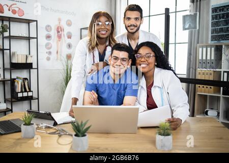 Gruppe von vier internationalen Ärzten in Laborkittel und -Peelings, die zusammen am Tisch sitzen und Laptop und Tablet benutzen. Medizinische Konferenz von männlichen und weiblichen Spezialisten. Mit Wissen teilen. Stockfoto