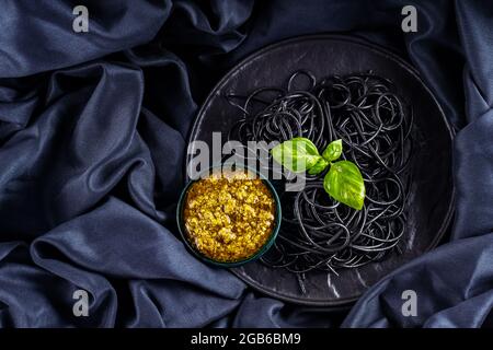 Schwarze Pasta, Pesto-Sauce und Basilikum auf dunklem Hintergrund. Schwarze Spaghetti mit Tintenfisch-Tinte auf einem schwarzen Tuch. Pasta auf schwarzem Teller und Sauce Pesto. C Stockfoto
