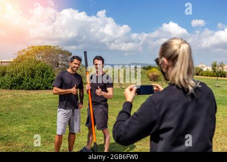Fitnesstrainer und Klientin posieren für eine Reporterin und schießen auf einem Smartphone. Sonniger Tag. Mit Gymnastikstab. Sommer im Garten. Stockfoto
