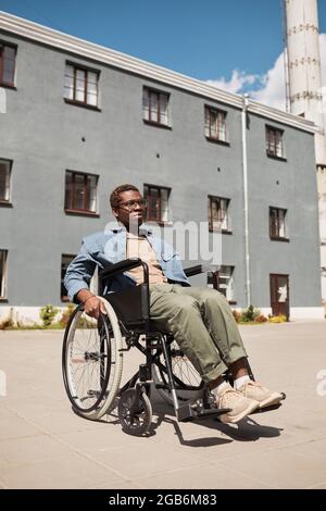 Ernst depressiver junger afroamerikanischer Mann in einer Brille, der im Rollstuhl gegen ein graues Gebäude sitzt und in die Ferne blickt Stockfoto