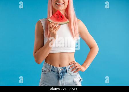 Ausschnitt einer jungen Frau mit rosafarbenem Haar, die eine Scheibe Wassermelone hält und lächelt und isoliert auf blauem Studiohintergrund posiert Stockfoto