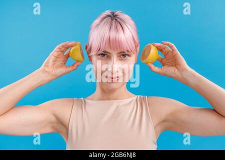 Porträt einer jungen Frau mit rosa Haaren, die lächelt und Hälften einer frischen Zitrone in der Nähe ihrer Ohren hält und isoliert auf blauem Hintergrund posiert Stockfoto