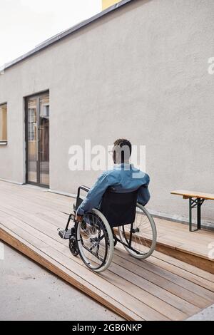 Rückansicht eines behinderten jungen afroamerikanischen Mannes, der im Rollstuhl sitzt und sich entlang einer Rampe in der Nähe des Hauses bewegt Stockfoto