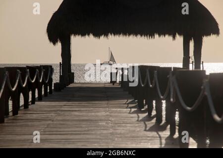 Hölzerner Pier auf dem Meer gegen das Licht mit einem Segelboot am Horizont, die tropische Insel Cozumel in Mexiko. Selektiver Fokus. Stockfoto