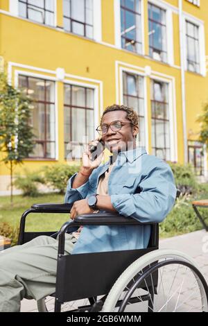 Porträt eines gut aussehenden jungen behinderten afroamerikanischen Mannes in Brille, der im Rollstuhl sitzt und per Telefon anruft Stockfoto