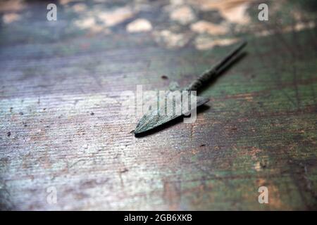 Nahaufnahme einer Pfeilspitze aus der Antike, aus Bronze, oxidierte Farbe auf einer ähnlich farbigen Holzoberfläche mit schwach grüner Farbe Stockfoto