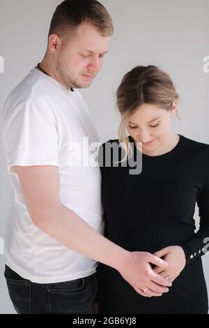 Die Schwangerin umarmt sich mit ihrem lieblichen Mann und legt die Hand auf den Bauch. Studio-Fotosession. Weißer Hintergrund Stockfoto