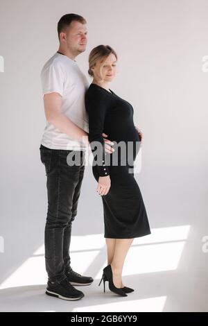 Die Schwangerin umarmt sich mit ihrem lieblichen Mann und legt die Hand auf den Bauch. Studio-Fotosession. Weißer Hintergrund Stockfoto