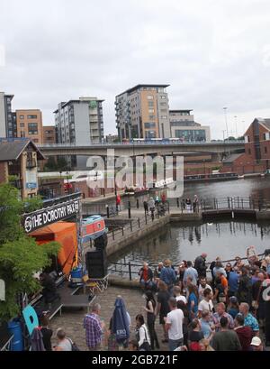 Sheffield England 25. Juli 2021. Eine Straßenbahn fährt in das Stadtzentrum von Sheffield über Barges, die an den Victoria Quays vertäut sind, während sich im Rahmen des Tramlines-Festivals eine Menge auf der Dorothy Pax-Bühne versammelt. Stockfoto