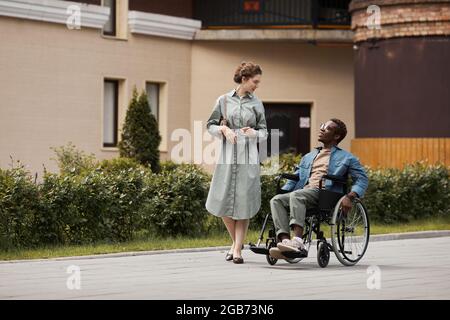Attraktive junge Frau in Kleid, die auf der Straße spazieren geht und mit einem behinderten Mann spricht, während sie sich mit ihm verquatet Stockfoto