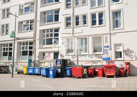 Eine Reihe farbiger Recycling-Mülleimer und Abfalleimer an einer weißen Wand, die teilweise mit Graffiti-Schrott-Etiketten bedeckt ist, England, Großbritannien Stockfoto