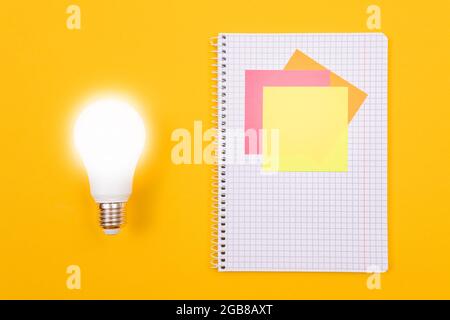 Ein Ideenkonzept - leuchtende Energiesparlampe und EIN Notizblatt mit Haftnotizen auf gelbem Tisch. Draufsicht, Flach Liegend, Leere Vorlage Stockfoto