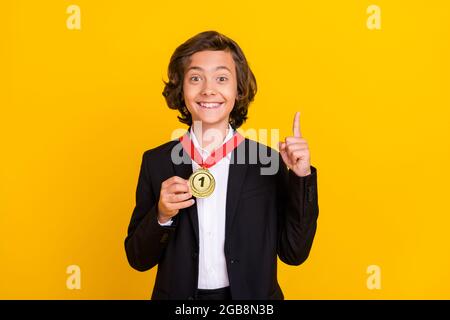 Foto des Genies Gewinner Junge zeigt ersten Platz Medaille erhöhen Finger tragen schwarze Uniform isoliert gelben Hintergrund Stockfoto