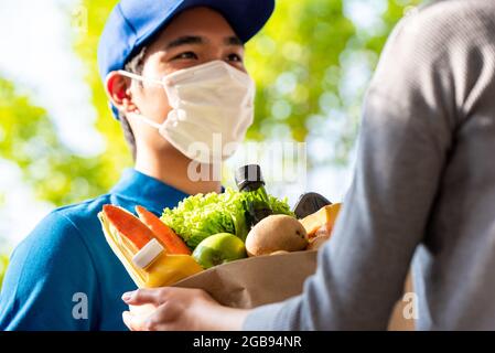 Hygienischer asiatischer Lieferer, der Gesichtsmaske trägt, während er Lebensmittel an den Kunden zu Hause liefert, und die Lieferung von Lebensmitteln in der Zeit des Pandemiekonzepts Stockfoto