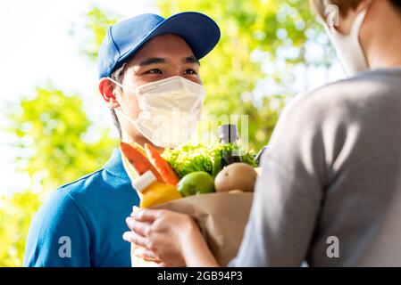 Hygienischer asiatischer Lieferer, der eine schützende Gesichtsmaske trägt, während er Lebensmittel an den Kunden zu Hause liefert, und die Lieferung von Lebensmitteln zur Zeit des Pandemiekonzepts Stockfoto