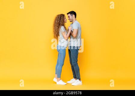 Portraitaufnahme eines jungen glücklichen, millenialen Paares, das in den Augen stand und sich in einem isolierten gelben Studio BA die Hände hielt Stockfoto