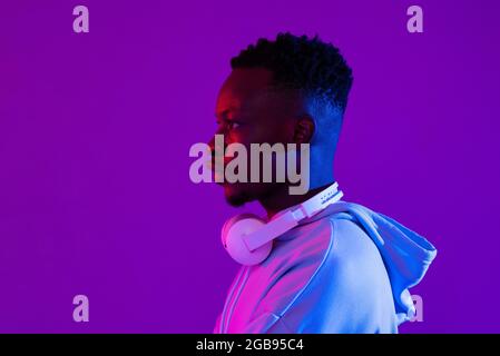 Seitenansicht Porträt von jungen schönen afrikanischen tausendjährigen Mann mit Kopfhörern in lila futuristischen Neonlicht Studio Hintergrund Stockfoto