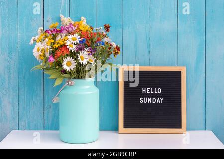 Happy Sunday Worte auf schwarzem Briefbrett und Strauß von hellen Wildblumen in Zinn können Vase auf Tisch gegen blaue Holzwand. Konzept Hallo Sonntag. Stockfoto