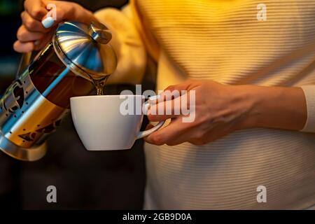 Die Hand eines Schwangeren hält einen weißen Becher, in den sie Tee aus einer schönen Teekannen gießt, um in einem dunklen Raum des Restaurants zu brauen Stockfoto