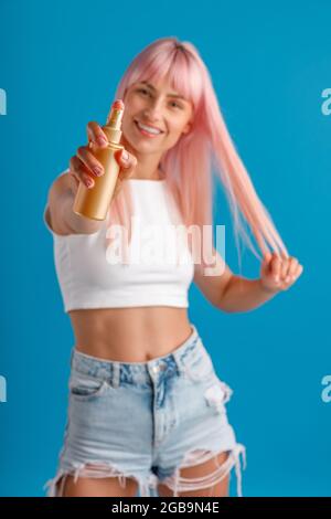 Fröhliche junge Frau mit rosa Haaren lächelt und zeigt feuchtigkeitsspendende Spray auf die Kamera, während sie isoliert über blauem Studiohintergrund steht Stockfoto