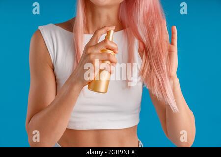 Nahaufnahme der Hände einer jungen Frau, die ihr glattes rosa Haar hält und feuchtigkeitsspendendes Spray auf sie anwendet, isoliert auf blauem Studiohintergrund stehend Stockfoto