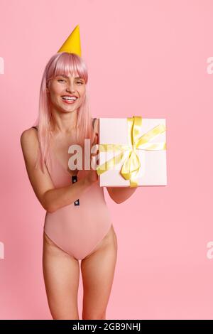 Lächelnde junge Frau mit rosa Haaren trägt Badeanzug und Partyhut mit niedlichen Geschenk-Box, isoliert auf rosa Hintergrund stehen Stockfoto