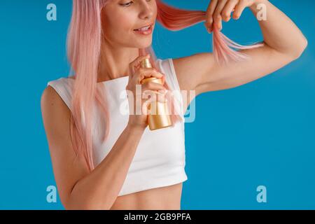 Nahaufnahme einer jungen Frau, die feuchtigkeitsspendende Sprays auf ihre rosa Haarenden anwendet, während sie isoliert auf blauem Studiohintergrund steht Stockfoto