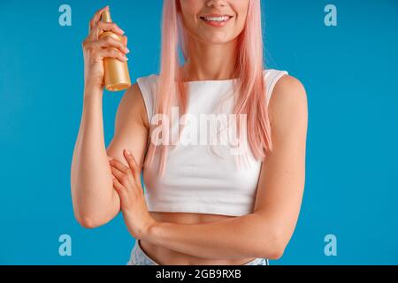 Kurzer Schuss einer jungen Frau mit rosa Haaren, die lächelt und eine Flasche feuchtigkeitsspendenden Sprays hält, während sie isoliert auf blauem Studiohintergrund steht Stockfoto